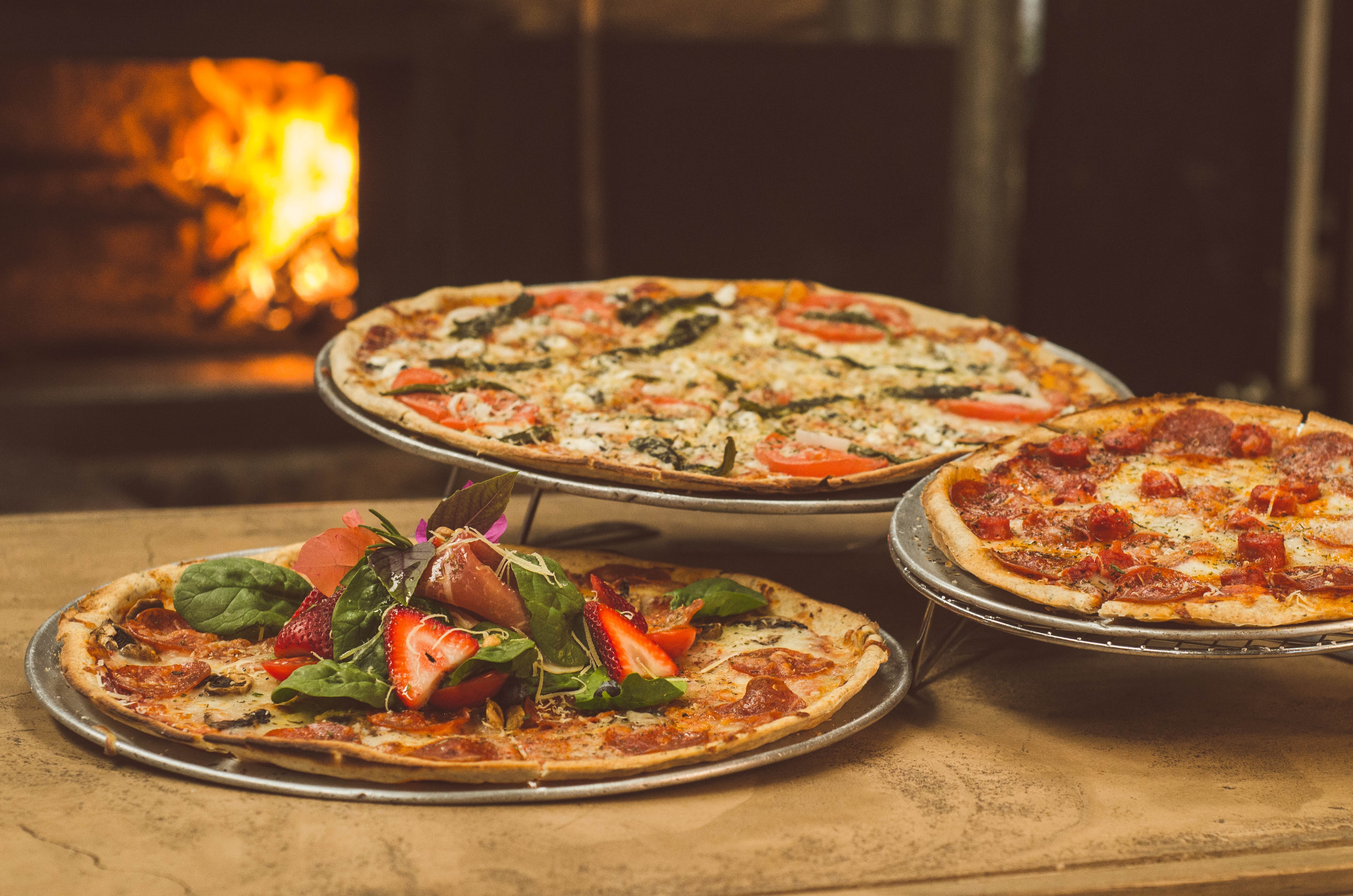 Three pizzas on pans in front of a fire place.
