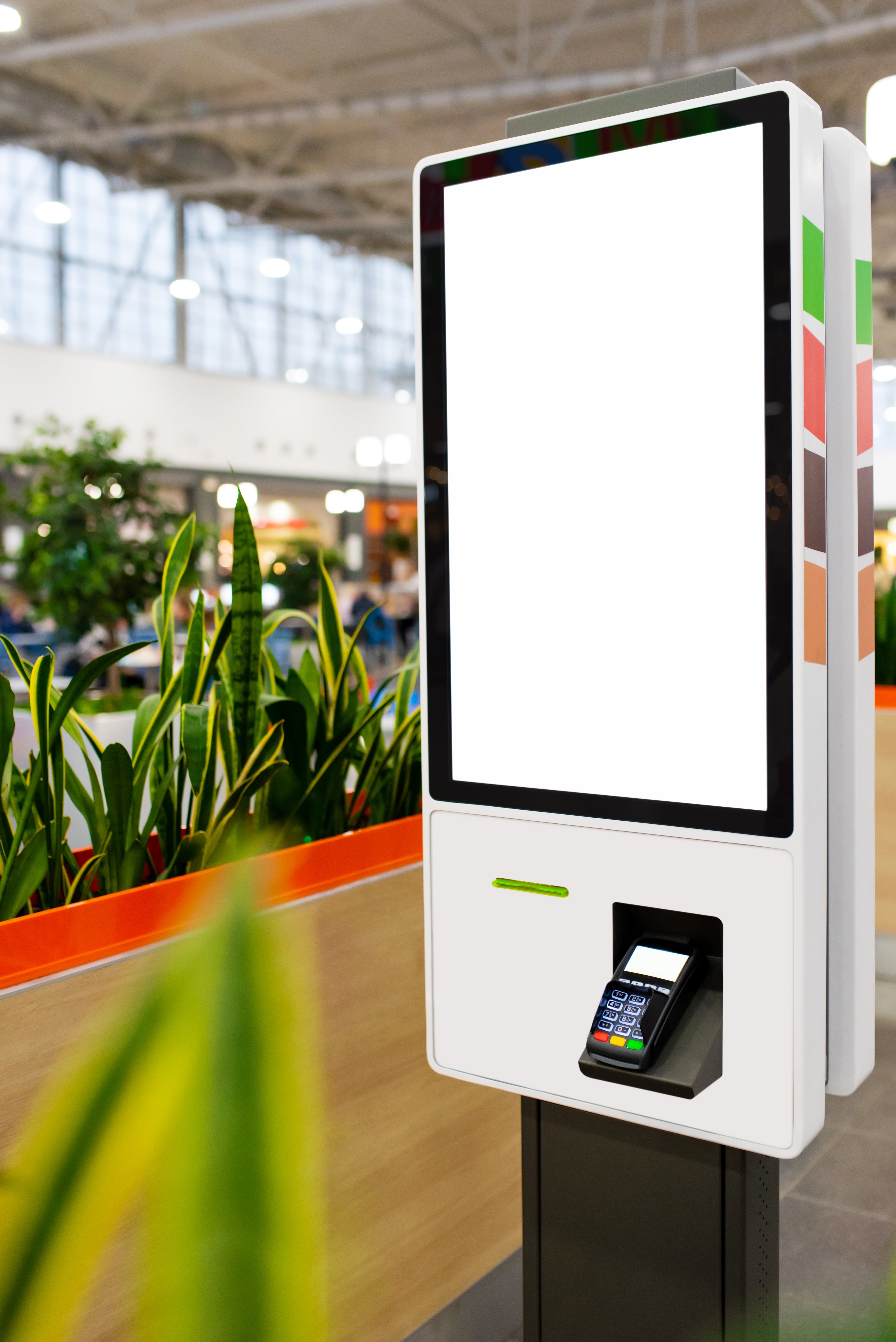 A white and green atm machine with plants in the background.